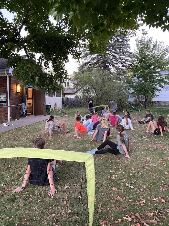 A group of children sitting on the grass in a backyard, with a person standing at the front, possibly leading an activity or game. There is a small soccer goal in the foreground, trees around the yard, and a house to the left.