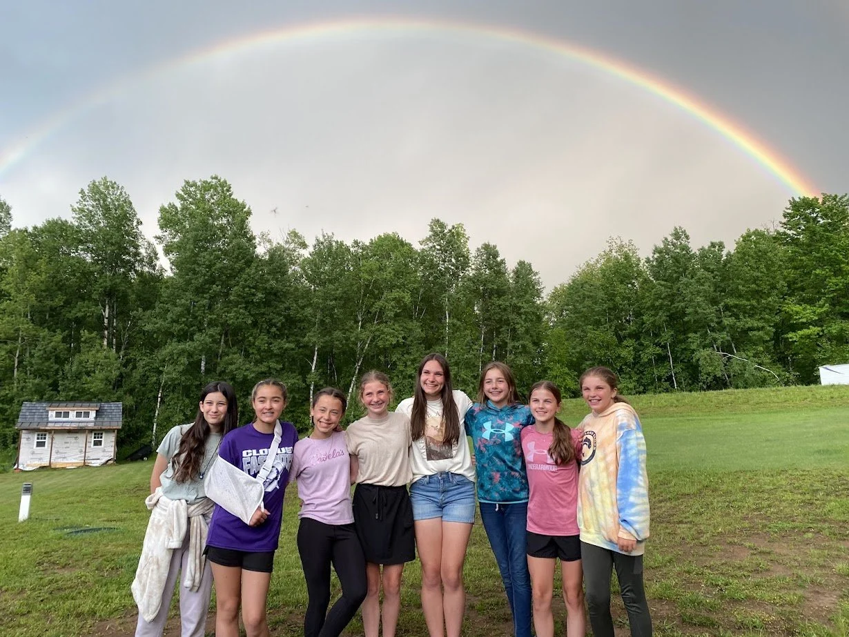 Eight girls smiling and standing together outdoors under a rainbow with a cloudy sky and green trees in the background.