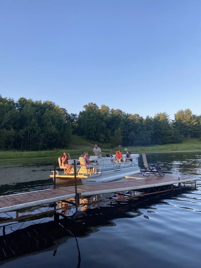 People preparing to board a pontoon boat at a dock on a lake, with a forested shoreline and clear blue sky in the background.