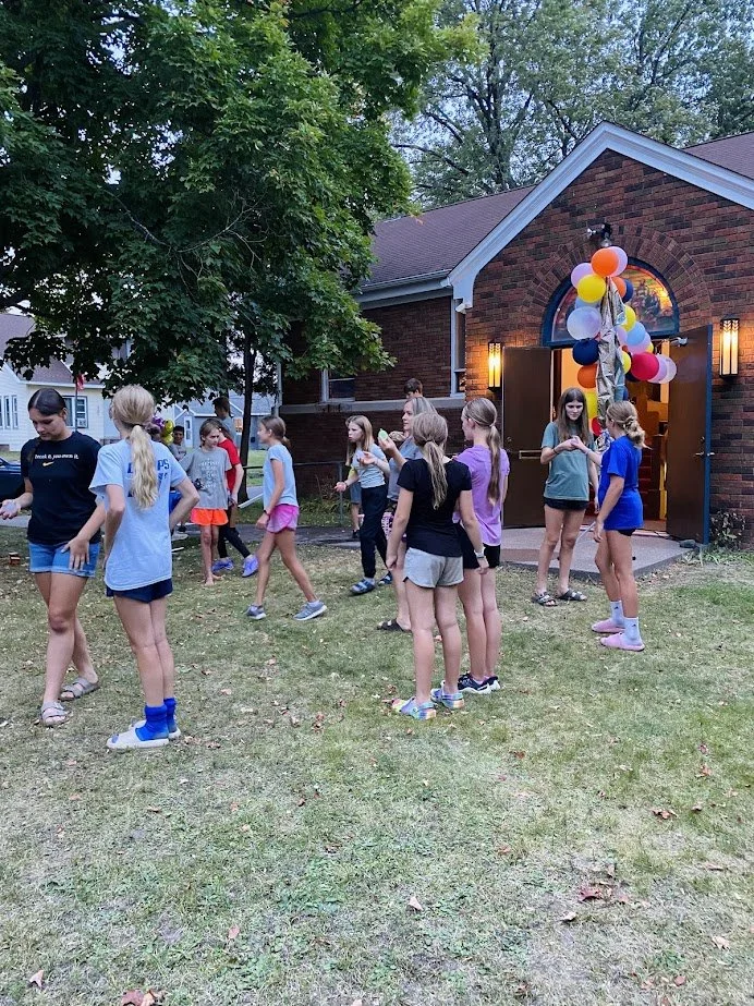 Group of children and teenagers gathered outdoors in front of a brick building decorated with balloons, likely for a celebration or party.