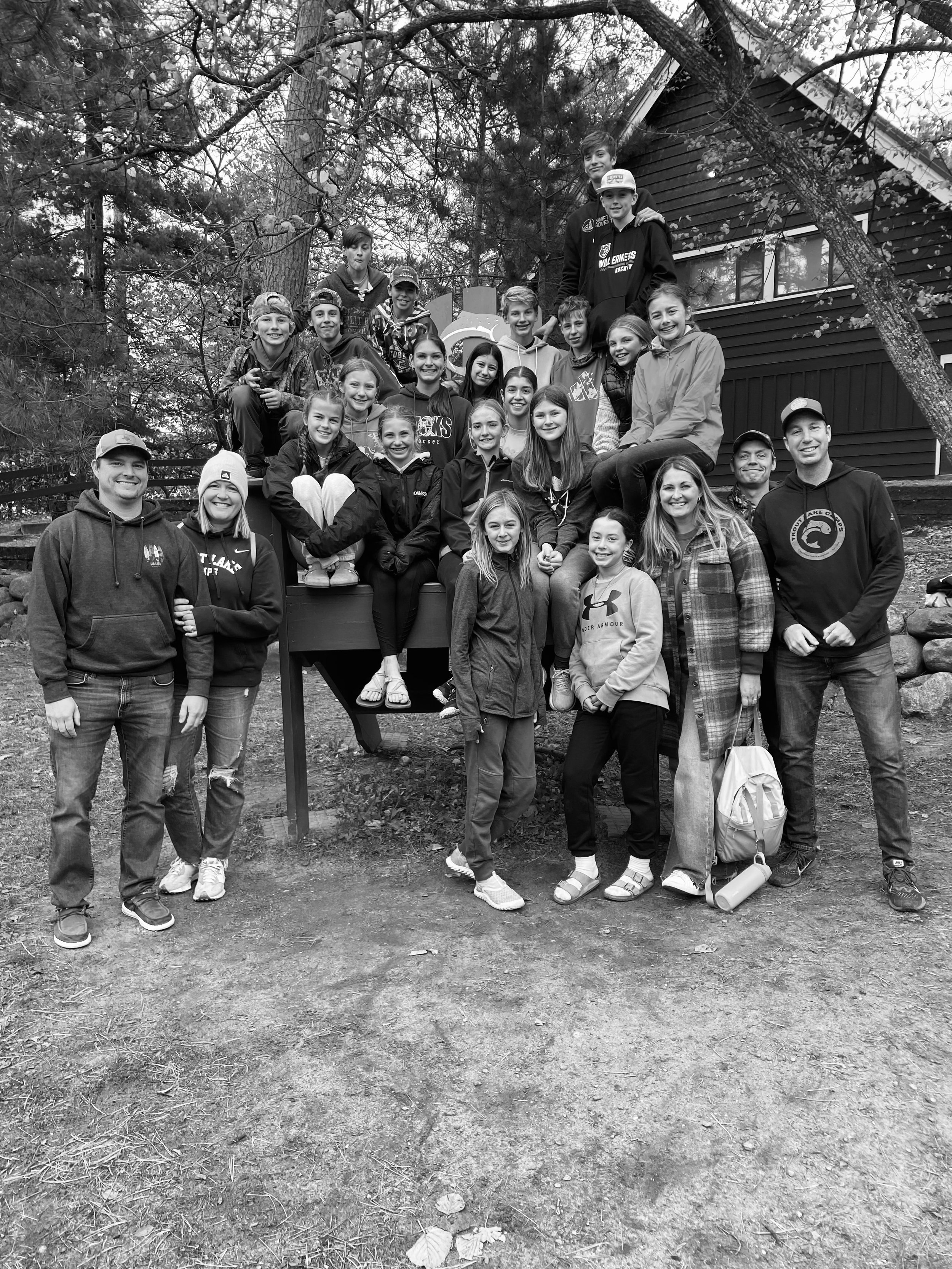 Group of people posing outdoors in front of a wooden building, surrounded by trees. Fall Retreat.