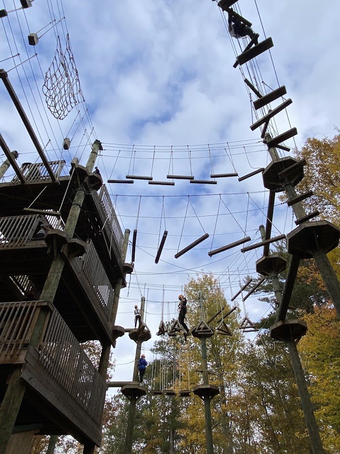Children and an adult walking on a treetop adventure course with wooden bridges, ropes, and platforms among trees with autumn leaves.