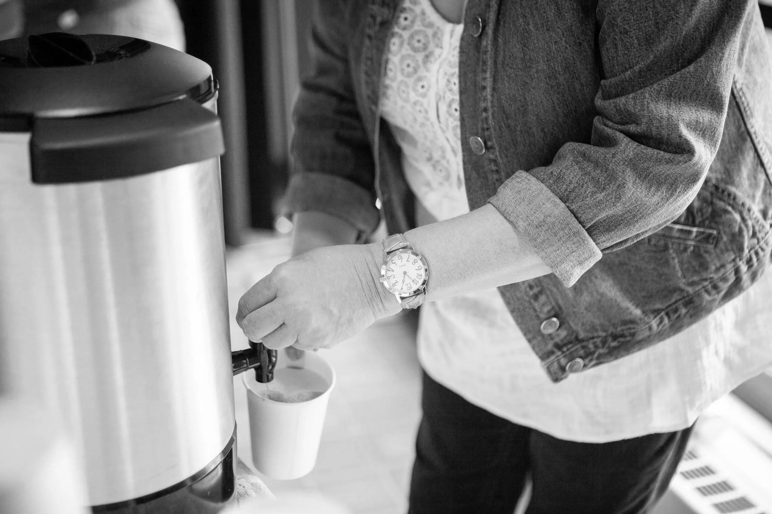 A person wearing a denim jacket and a watch is filling a cup from a coffee machine.