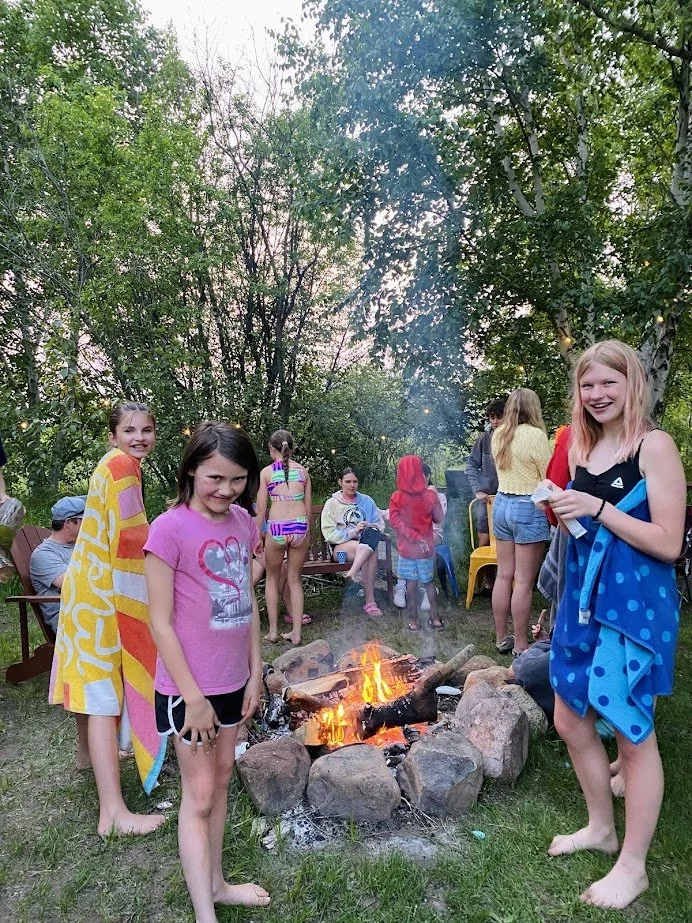 Children gathered around a campfire in a wooded area, some wrapped in towels, during a outdoor gathering or celebration.