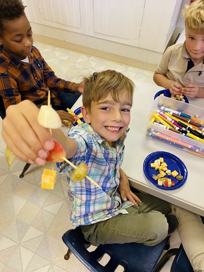 Boy smiling and showing off a fruit skewer at a table with two other children and art supplies.