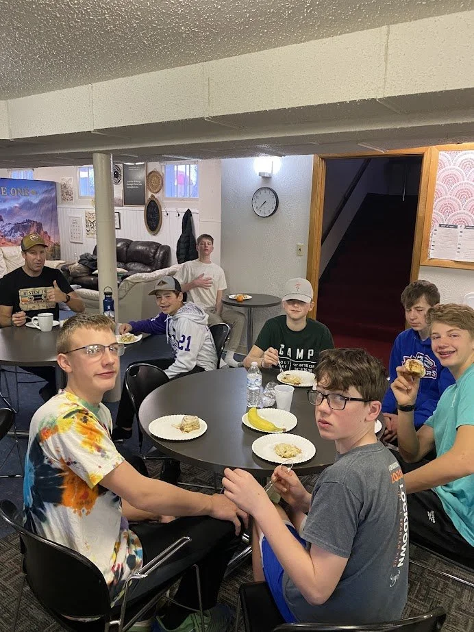 Group of boys sitting around tables in a room, eating snacks and drinking water, with some holding food and others with plates of dessert.