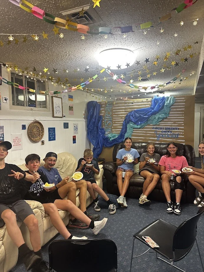 Group of children sitting on couches and chairs in a decorated room, enjoying snacks. The room has streamers and stars hanging from the ceiling, with a backdrop featuring fabric and a sign that says 'Discipleship Community'.
