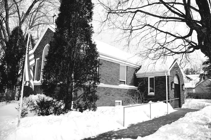 Brick building with a cross, surrounded by snow, trees, and an American flag.
