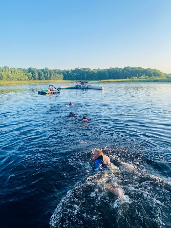 People swimming in a lake near a floating platform with a water slide on a sunny day, with trees in the background.