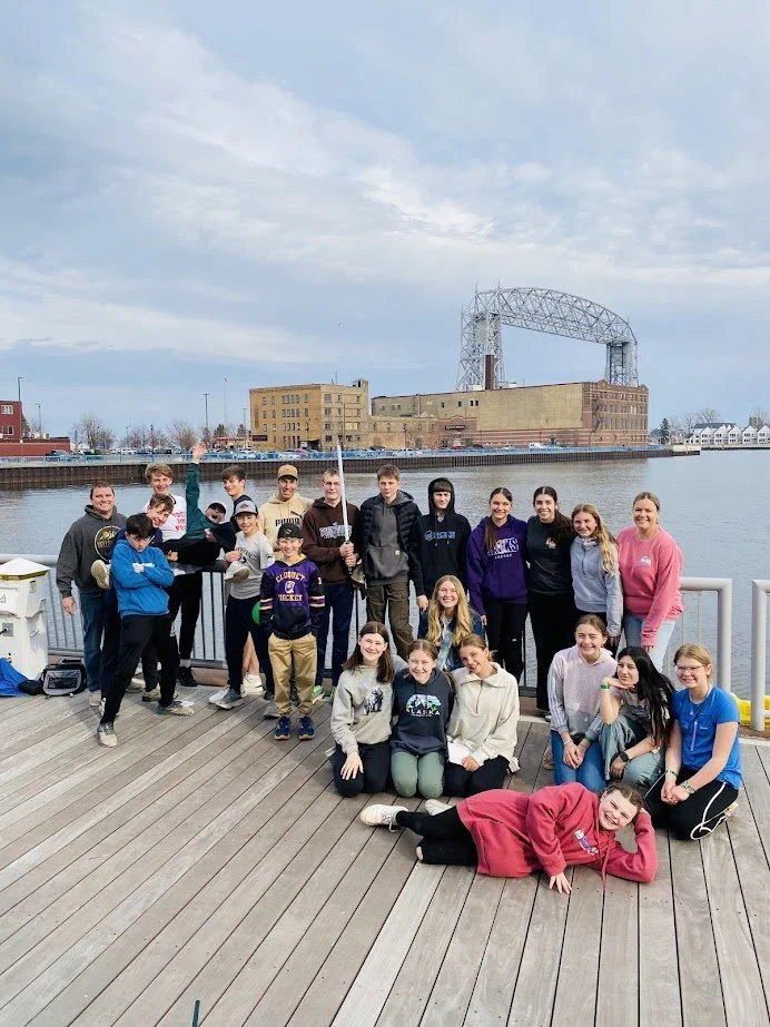 A group of teenagers and young adults posing on a wooden dock near a body of water with industrial buildings and a bridge in the background.
