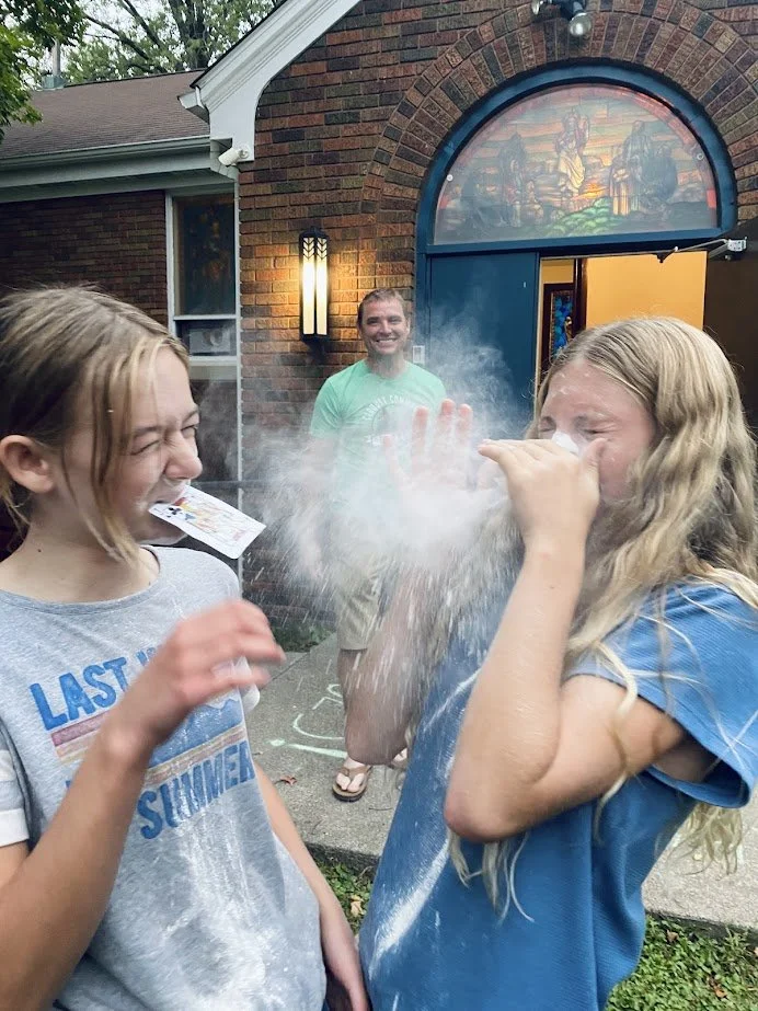 Two young girls outdoors, one sneezing into her hand with powdered sugar on her face, while the other girl laughs with powder on her face and shirt. A man stands in the background smiling, near a brick house with a stained glass window, at dusk.