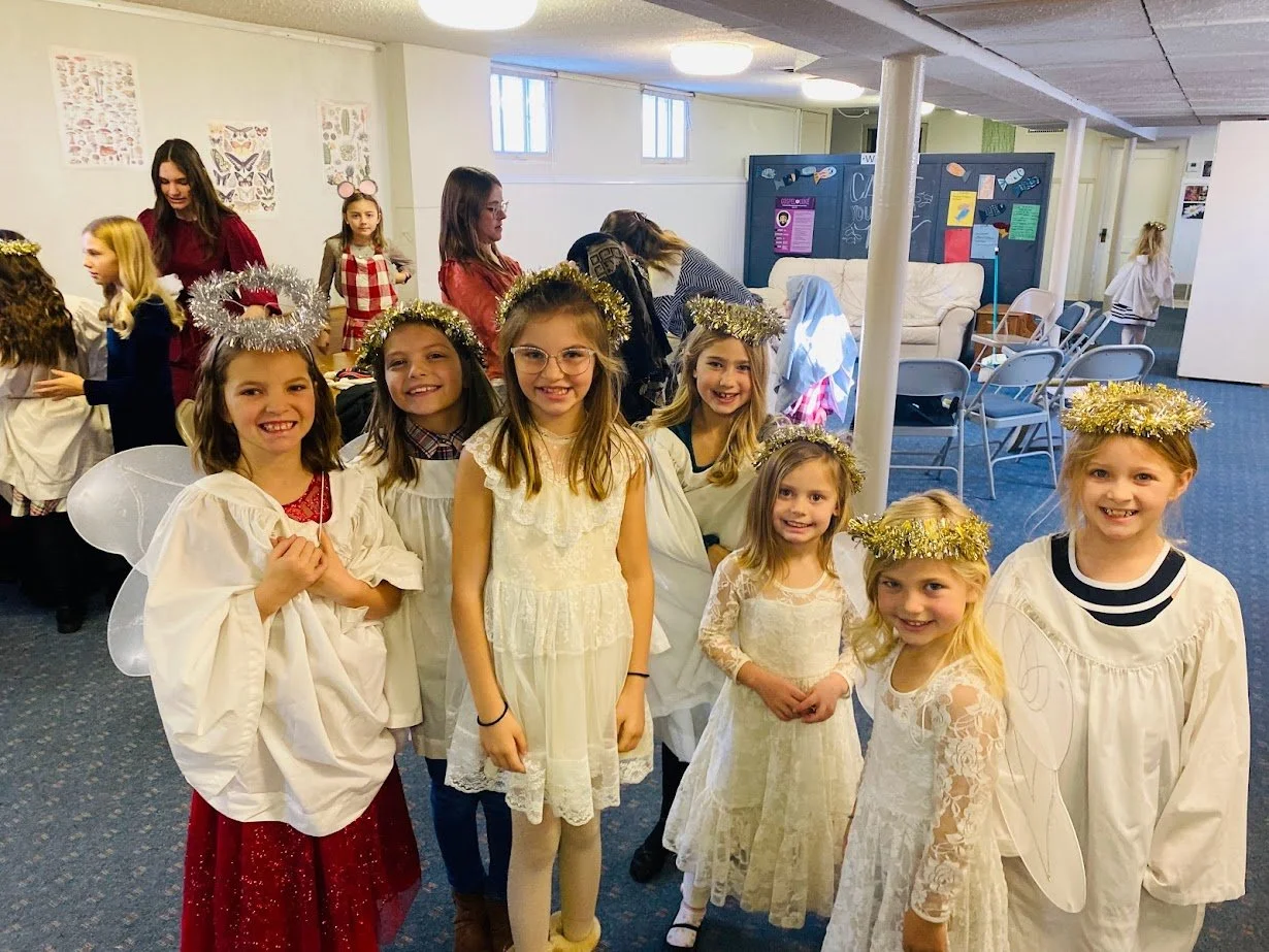 Group of young girls dressed as angels, wearing white dresses and golden tinsel halos, standing inside a decorated room, possibly for a holiday or Christmas event.