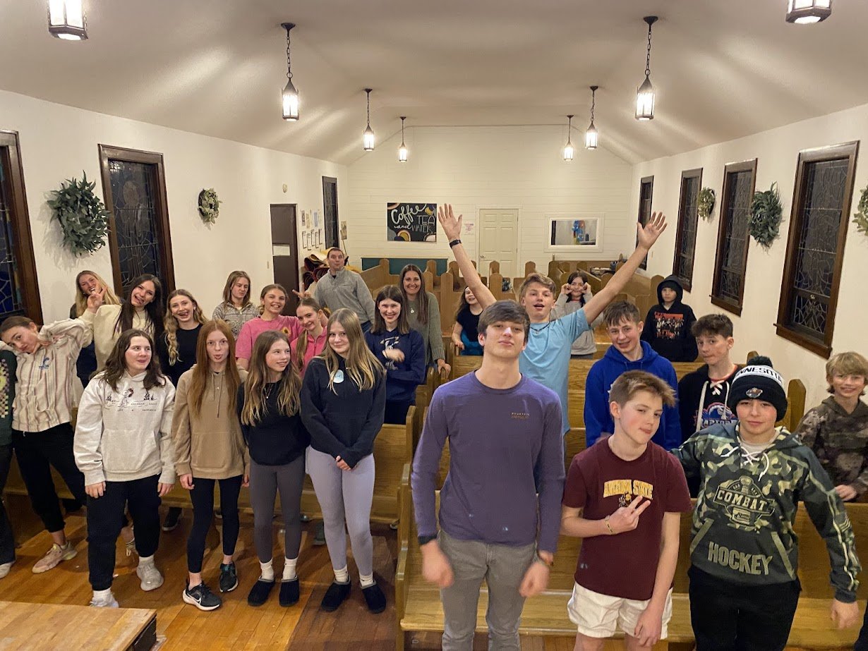 A group of teenagers inside a warmly lit room, posing for a photo with some smiling, others making playful gestures, and one person in the back raising their arms.