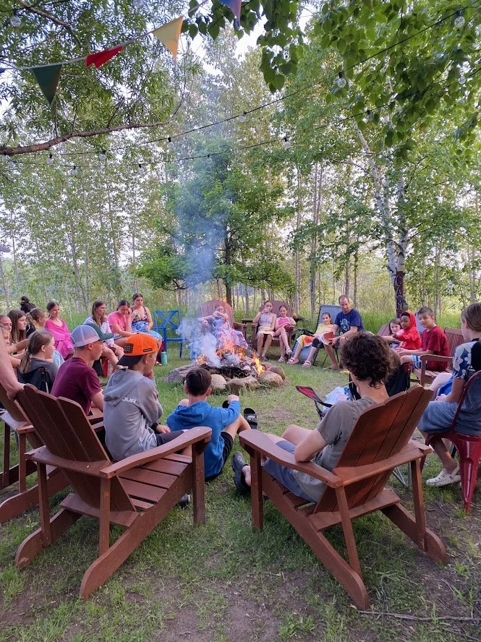 Group of children and adults sitting in a circle around a campfire outdoors, with trees and string lights overhead, possibly during a camping event or gathering.