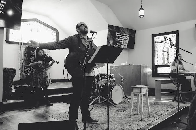 Black and white photo of a church worship band performing. A guitarist and singer stands at a microphone with a music stand, a keyboard player is seated on the right, and a violinist on the left. A drum set is visible in the background. Stained glass windows and a screen with text are in the background.