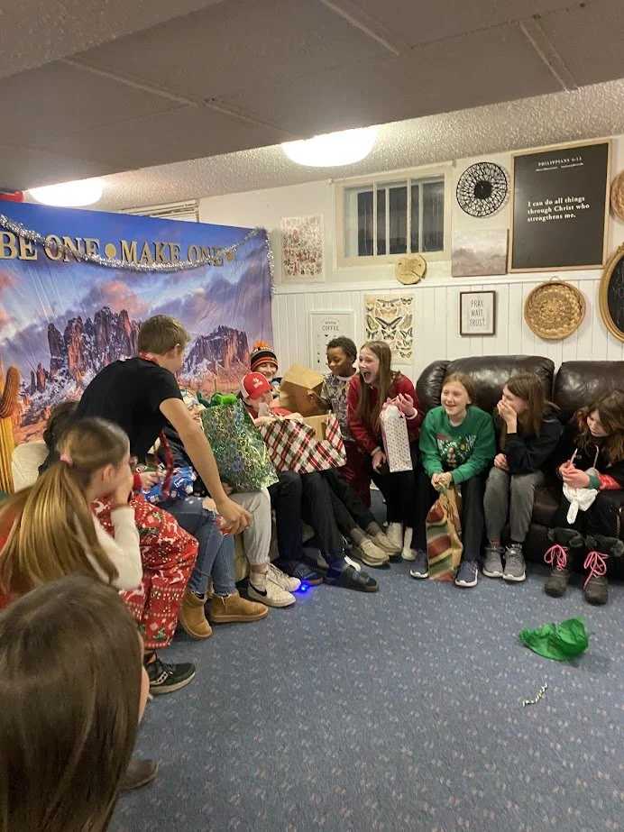 Children gathered in a living room for a gift exchange, with some opening presents and others watching and laughing.