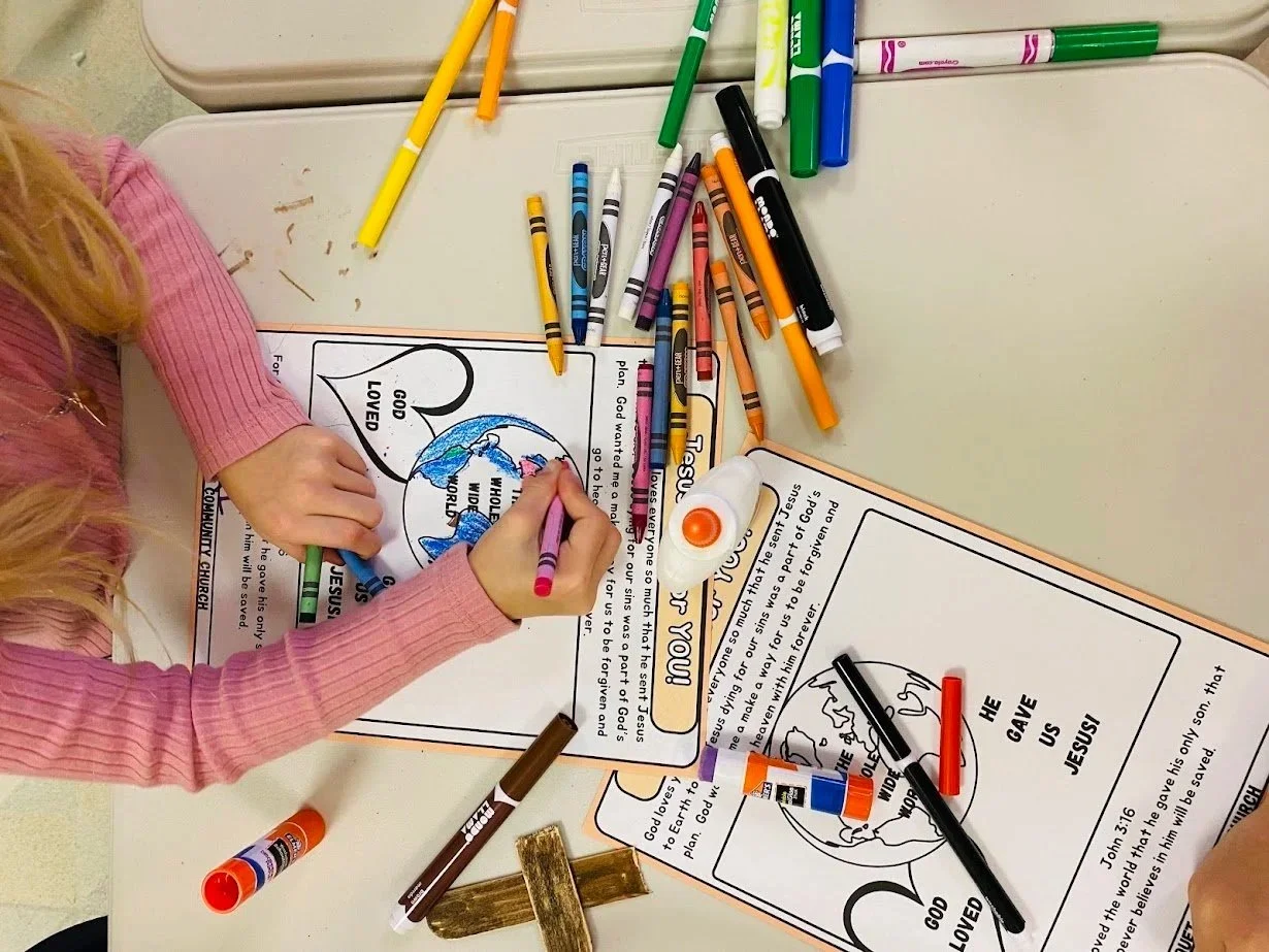 Two children coloring Easter-themed drawings with markers on a table outdoors, with a toy rabbit and a yellow lantern in the background.