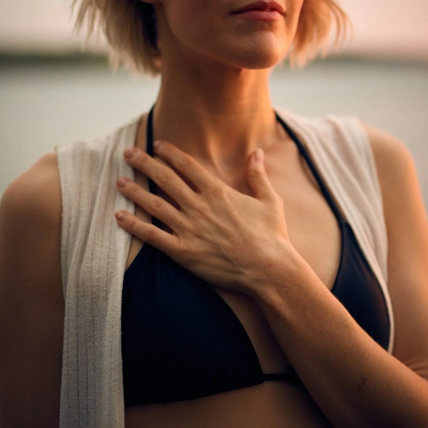 A woman with short hair wearing a black sports bra and a white sleeveless outer shirt, holding her hand on her throat in a gesture of distress or concern.