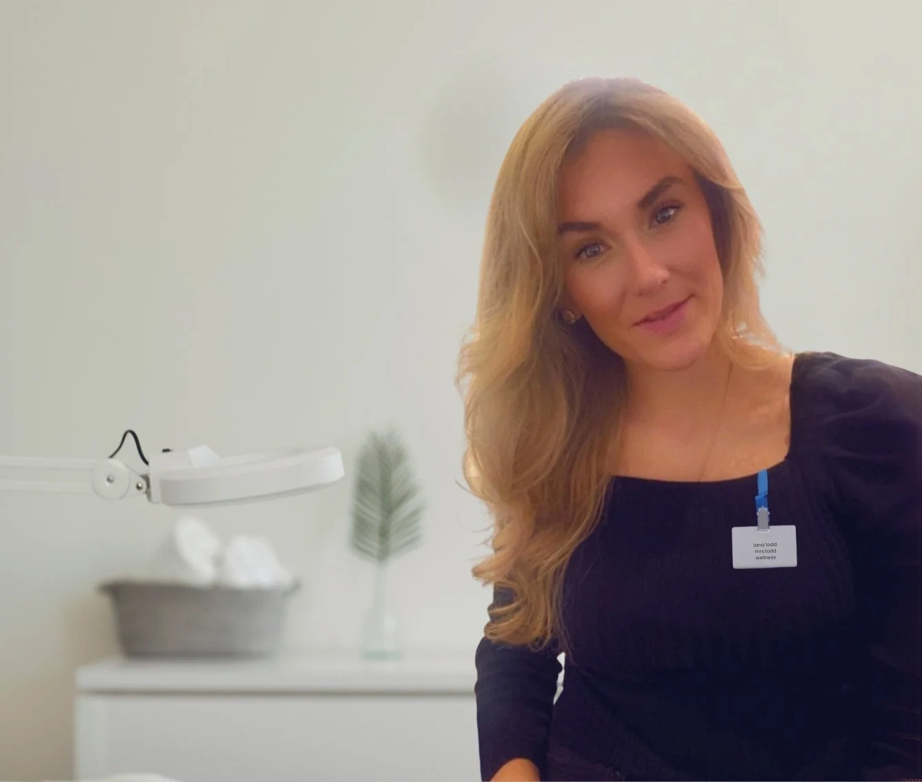 A woman with long, wavy blonde hair wearing a black top and a name badge sitting in a wellness or medical office. There is a white lamp, a basket with tissues, and a small vase with a green leaf in the background.