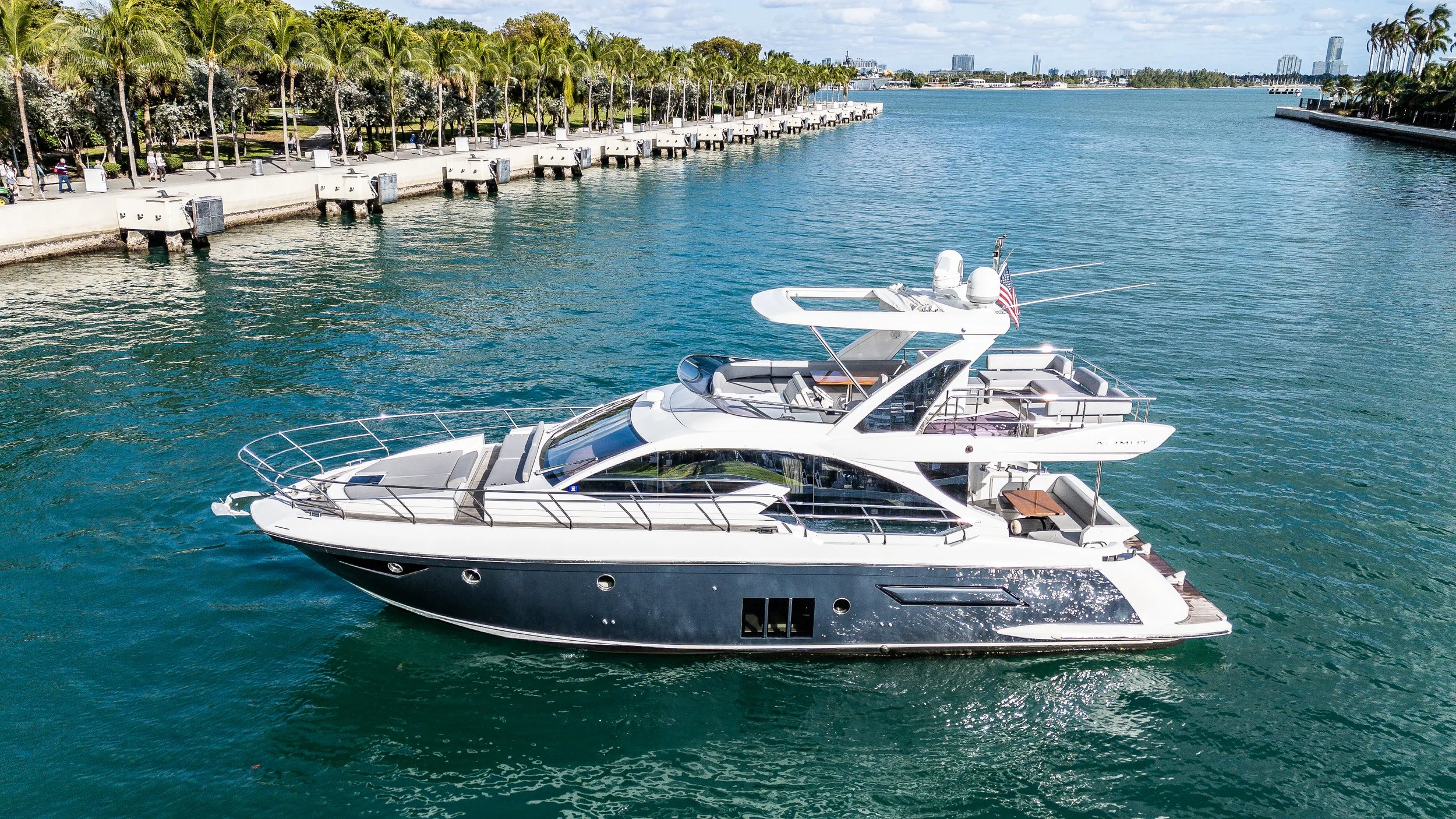 A luxury yacht floating in a marina near a landscaped waterfront with palm trees and city buildings in the background.