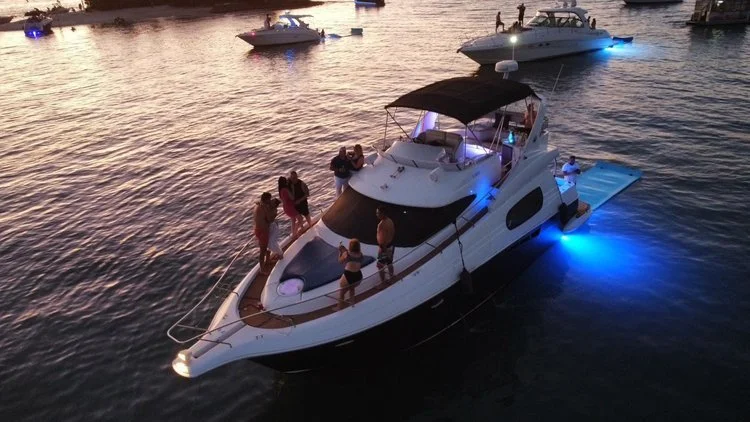 People relaxing on a white yacht with blue underwater lights, docked in a marina during sunset.
