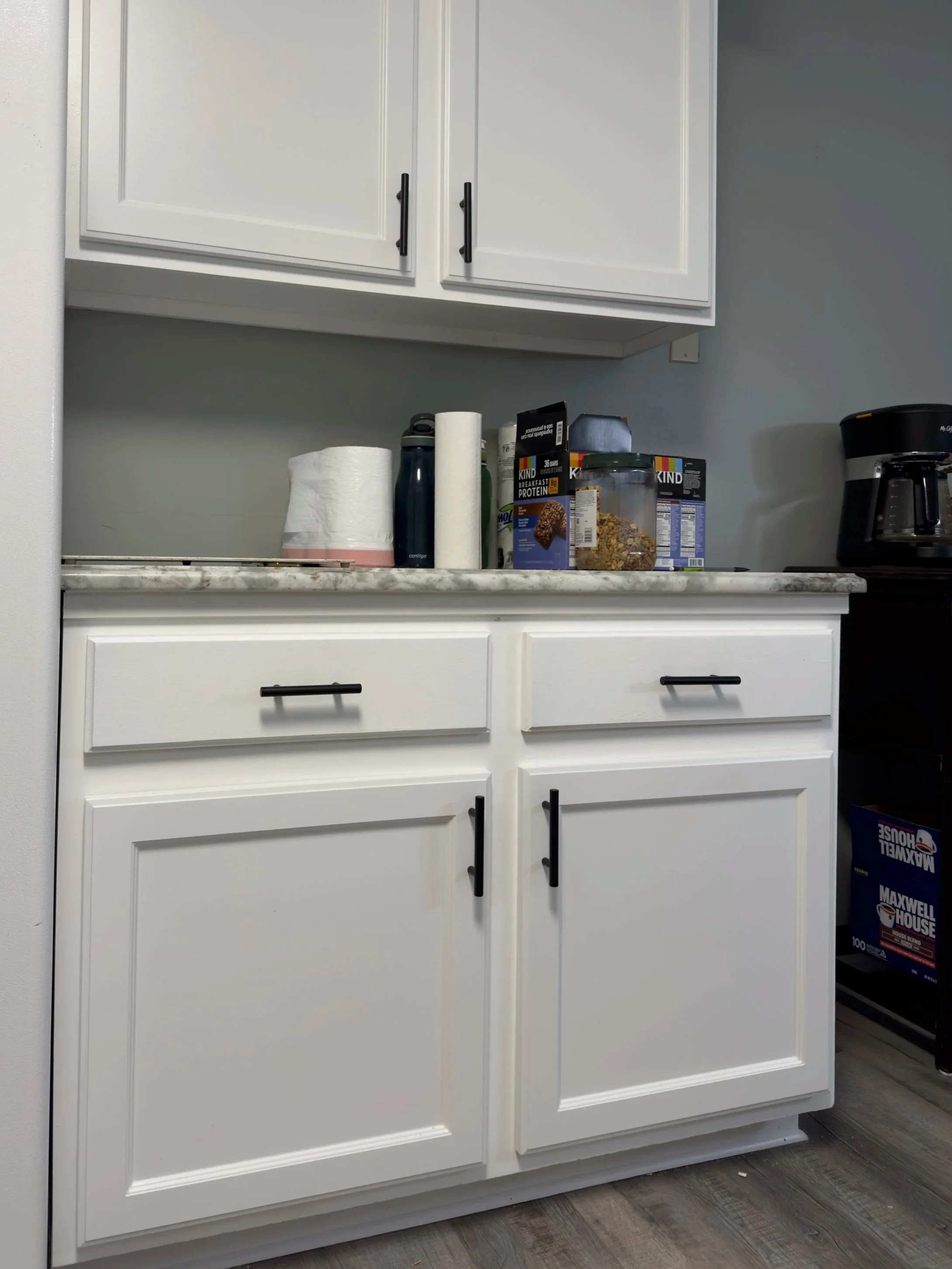 Kitchen countertop with paper towels, water bottle, cereal boxes, and a bottle of syrup. White cabinets beneath and above the counter. A coffee maker on the right.