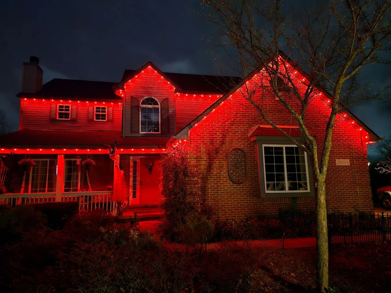 A brick house decorated with red Christmas lights at night, with a tree in the front yard and a dark sky in the background.