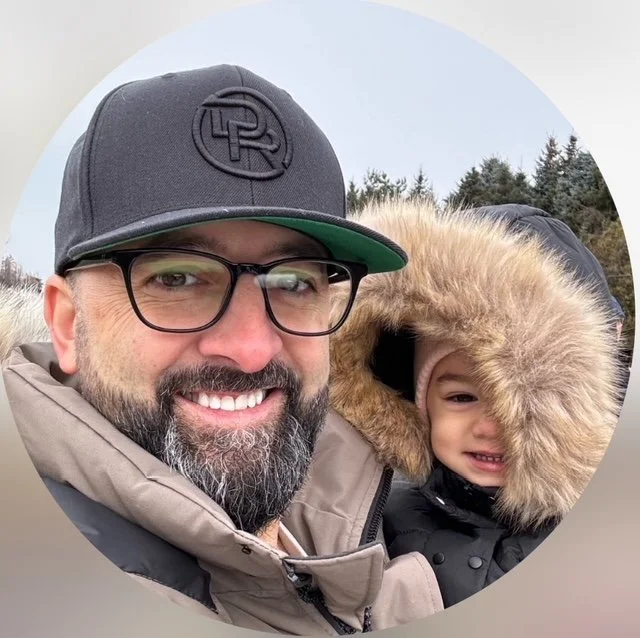 Man wearing glasses and a black cap, smiling with a child bundled in a fur-lined hood outdoors, with evergreen trees in the background.