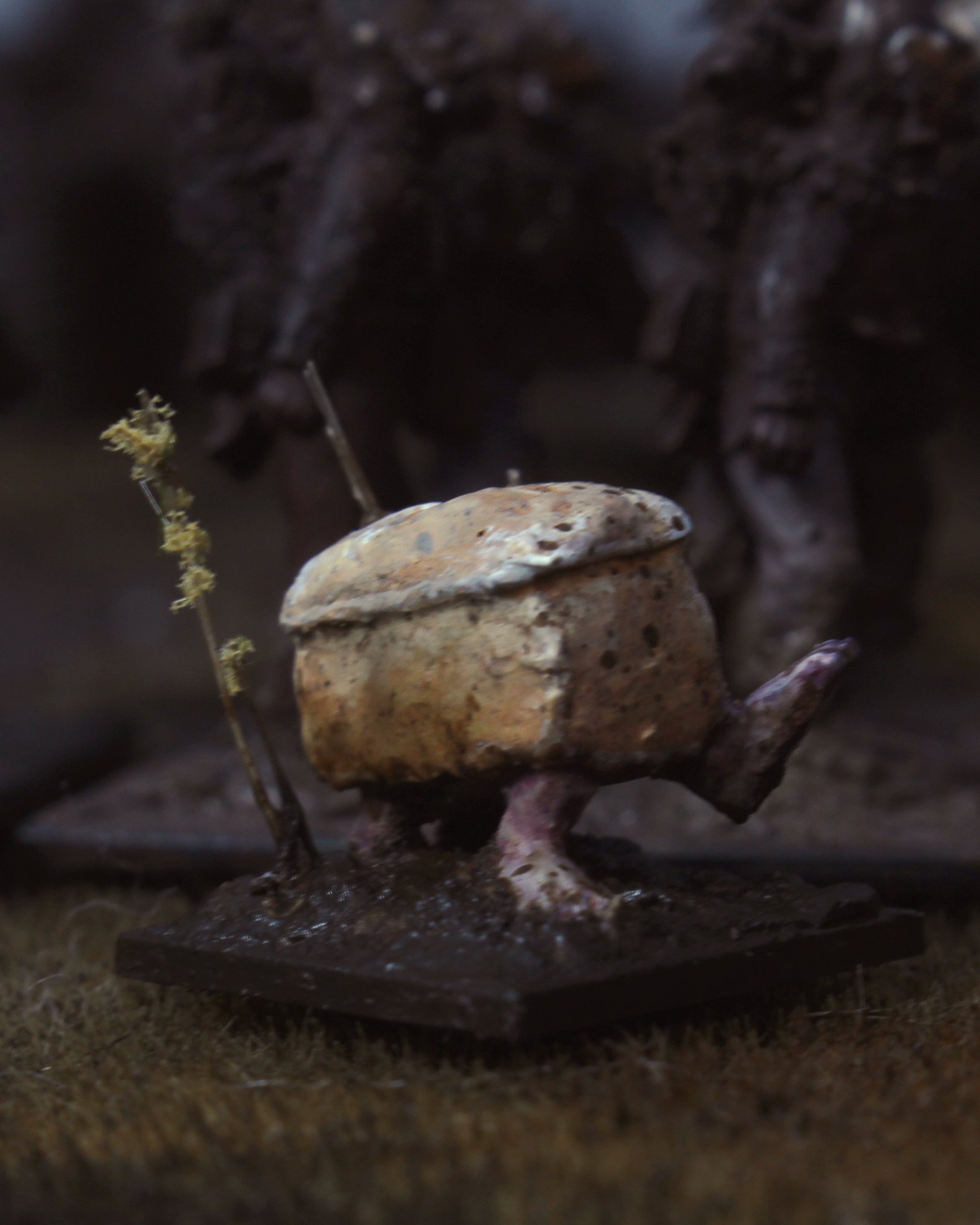 A small sculpture resembling a bird with a mushroom-shaped head and body, standing on a rectangular base with thin legs, with a background of dark figures and a tiny dried plant near the sculpture.