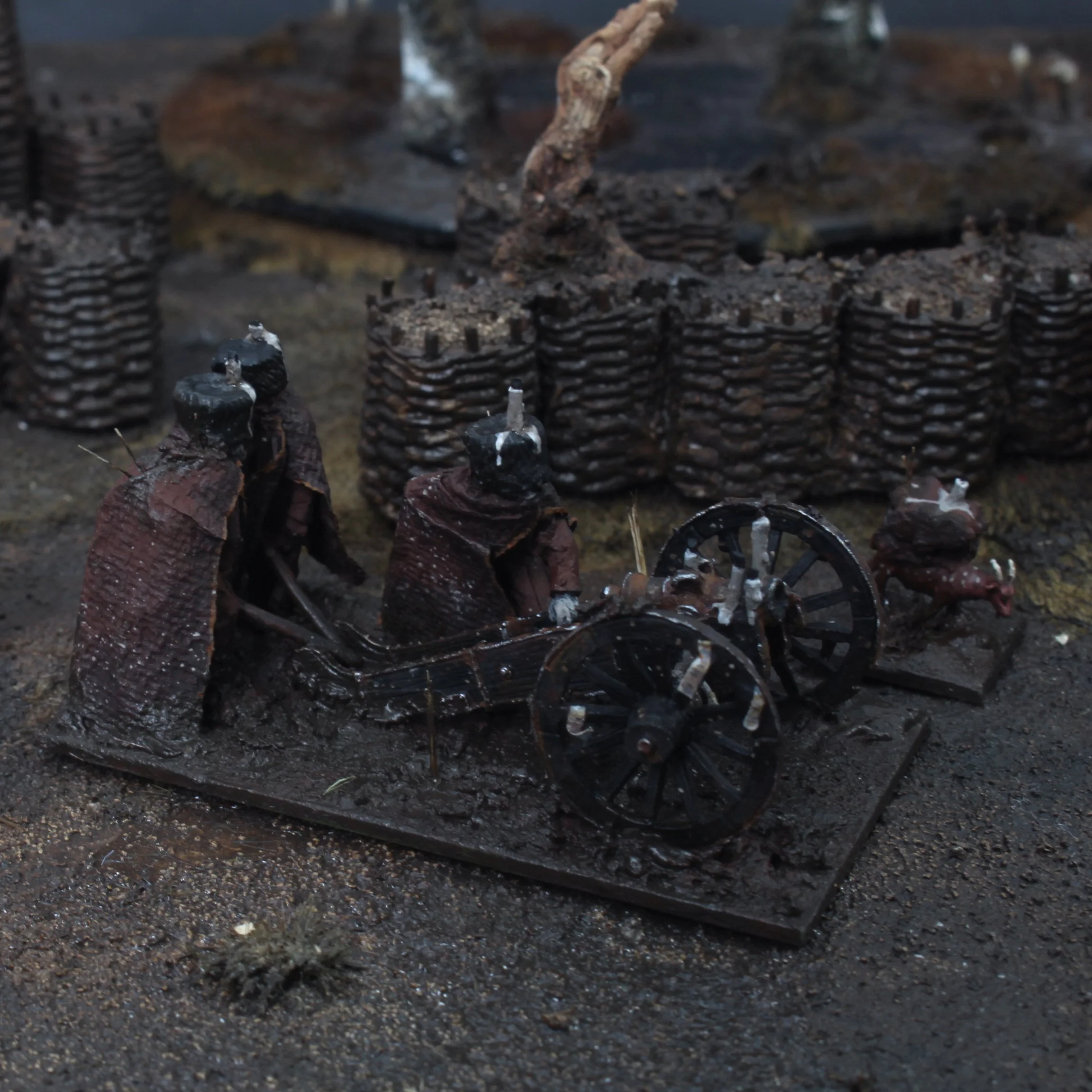 Small model of a cannon with three figures in dark clothing and hats, surrounded by stacks of black cylindrical objects, on a dirt surface.