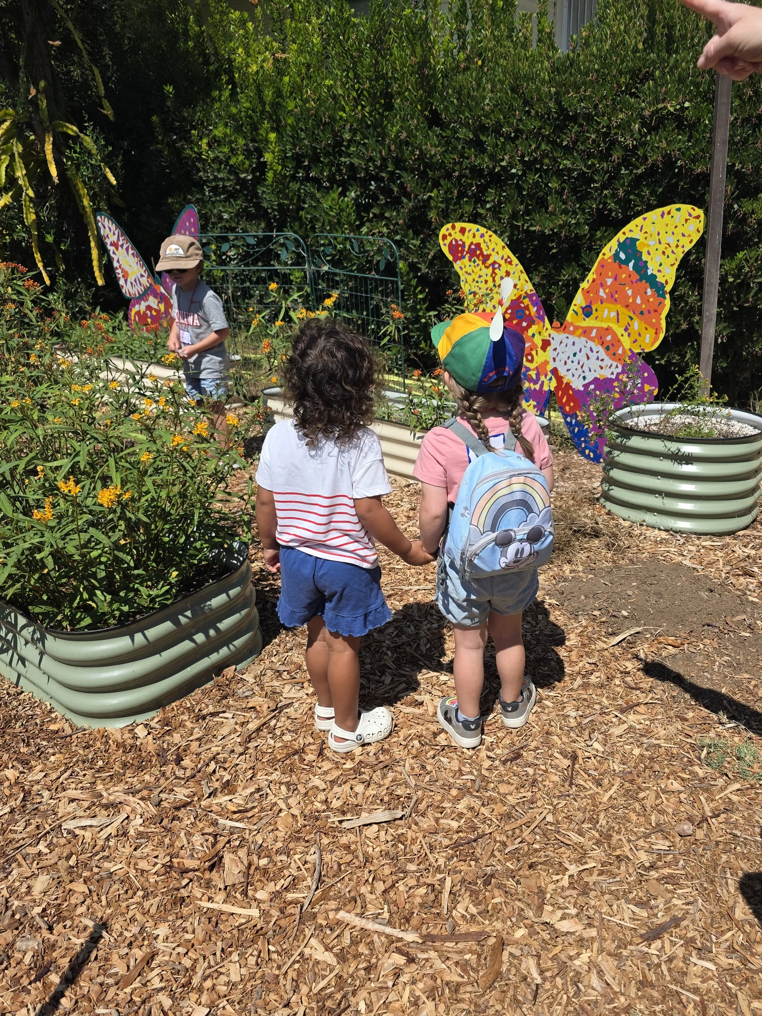 Four children standing on wood chips in a garden, holding hands, with colorful butterfly decorations in the background, and lush green bushes and trees surrounding them.