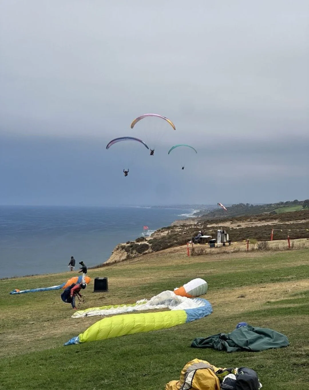 Paraglider pilots preparing on a grassy hilltop with three pilots already airborne over a coastal landscape.