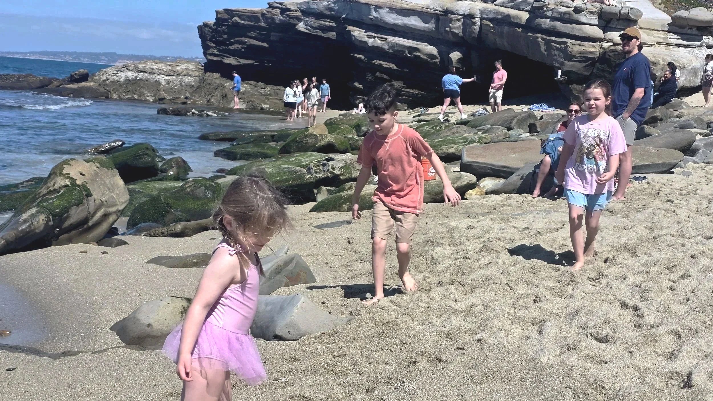 Children playing on a sandy beach with rocks, a large rock formation in the background, and several people walking and taking photos near the water and rocks.