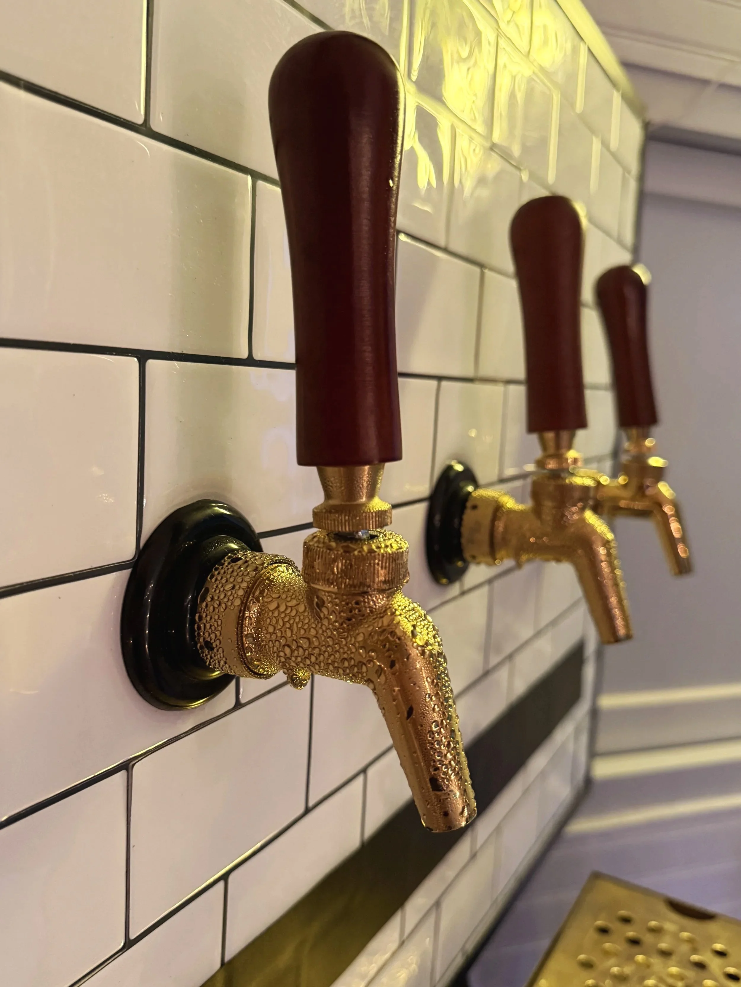 Close-up of beer taps with dark wood handles and gold fixtures mounted on a white tiled wall.