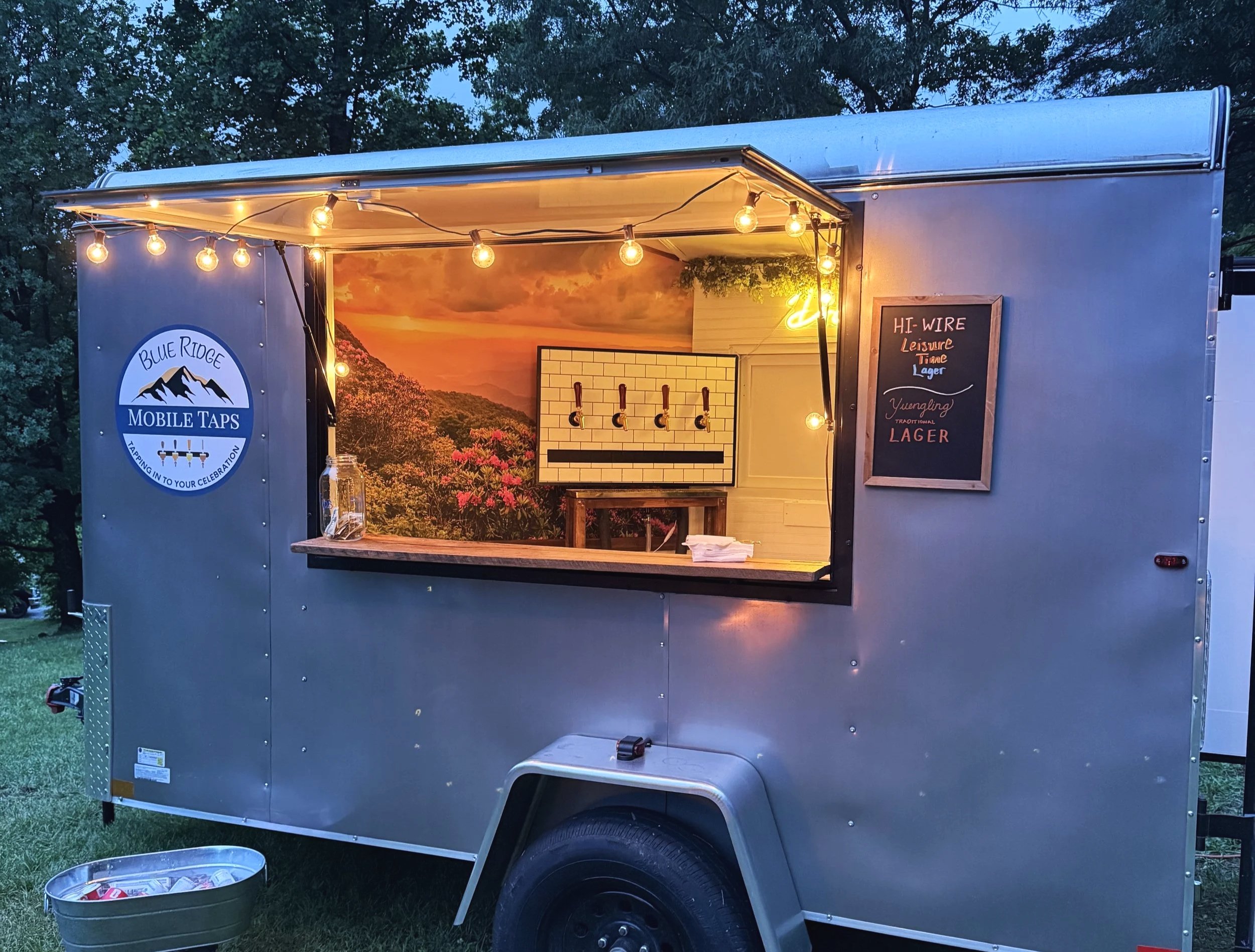 A small mobile tap trailer with string lights hanging from the top and a scenic mountain and sunset mural inside. There is a blackboard sign reading 'HI-WIRE Leisure Time Lager' and a logo for Blue Ridge Mobile Taps on the exterior. The trailer is parked outdoors on grass with trees in the background.