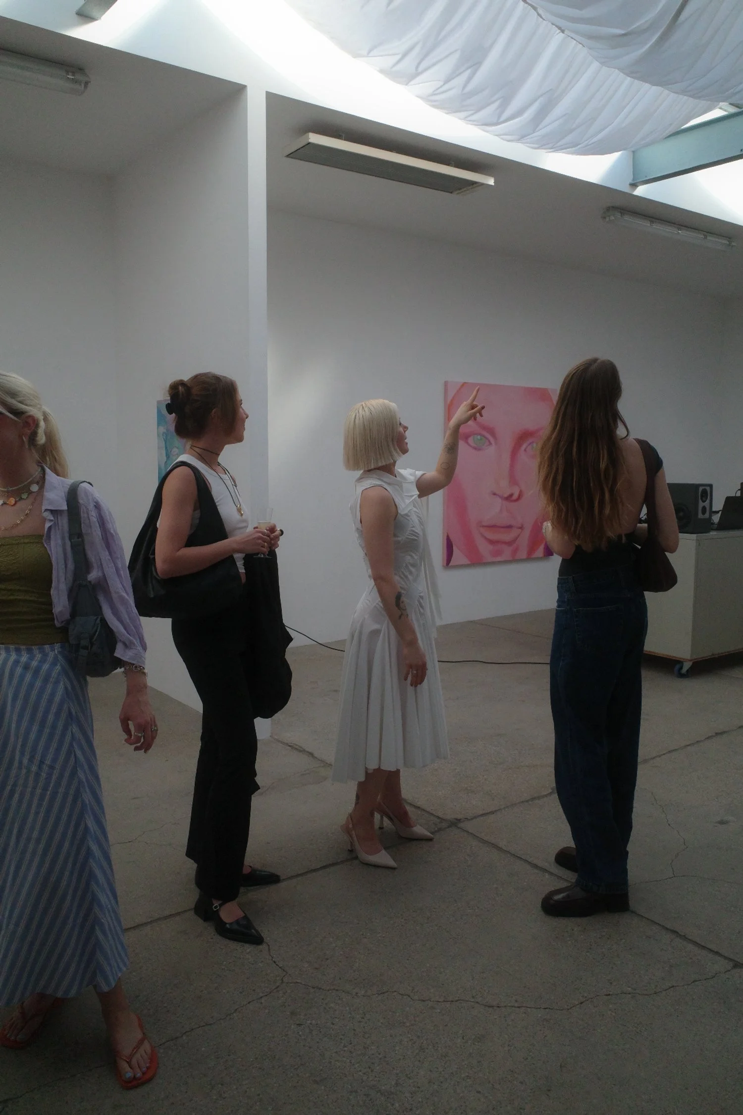 Menschen in einer Kunstgalerie, eine Frau spricht und zeigt auf ein Gemälde eines Gesichtes in Pink, andere stehen in einer Reihe und schauen zu.