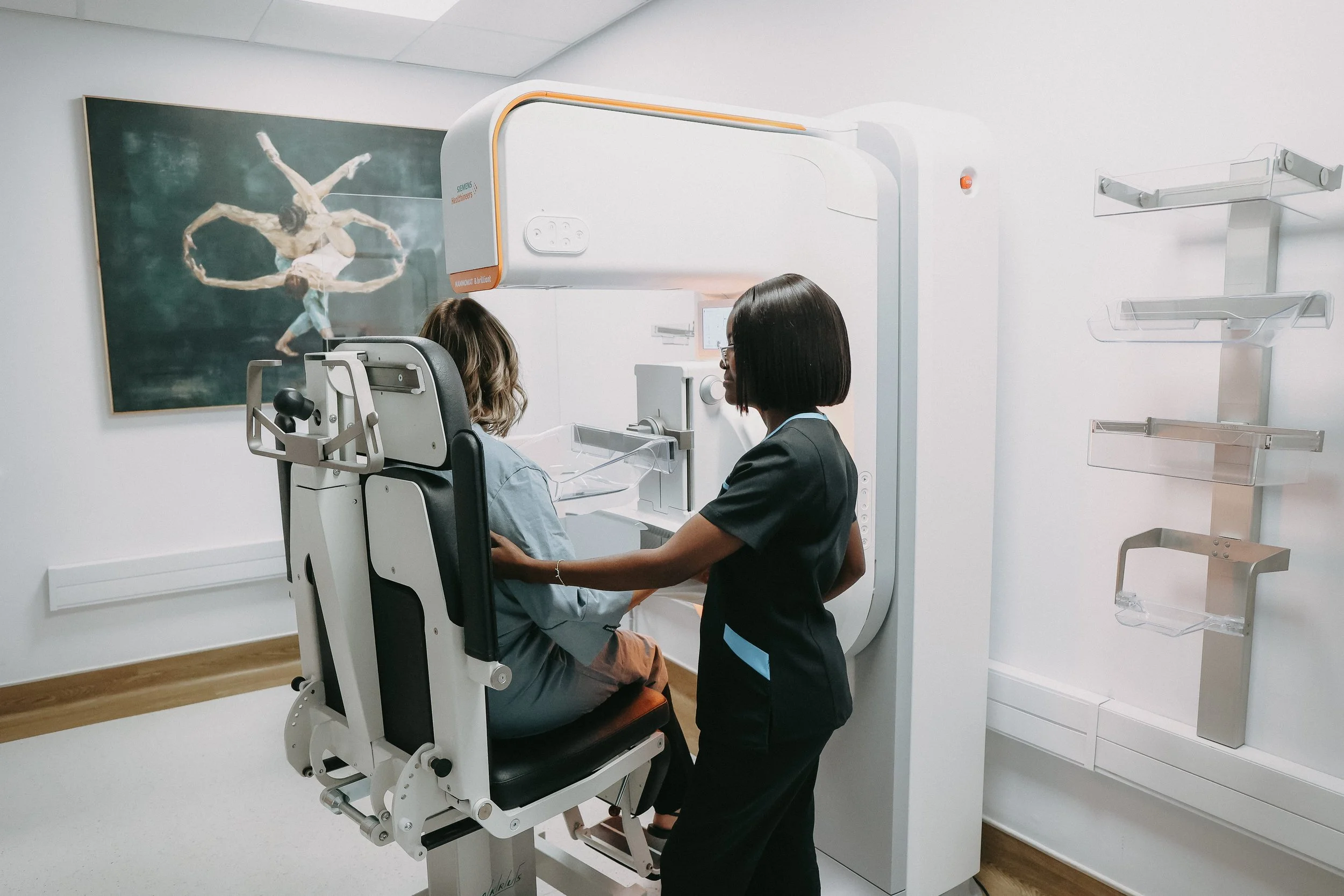 A woman getting a mammogram at the Light Room at NAMRAD while a technician points at a monitor displaying a breast image in a medical office.