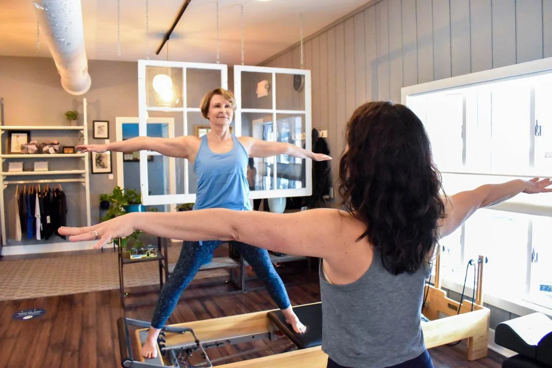 Two women are practicing yoga on Pilates reformers in a bright, cozy room with wooden floors and light-colored walls.