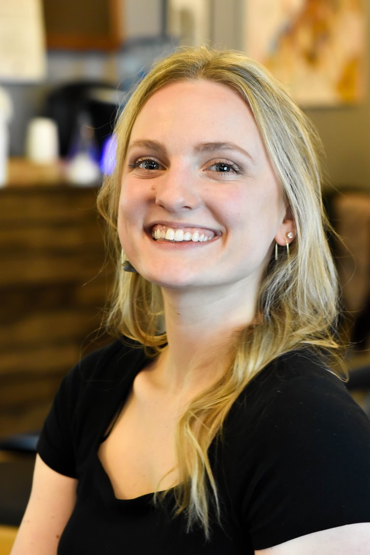 A young woman with long blonde hair, smiling, wearing a black shirt, in an indoor setting with a wooden counter and blurred background.