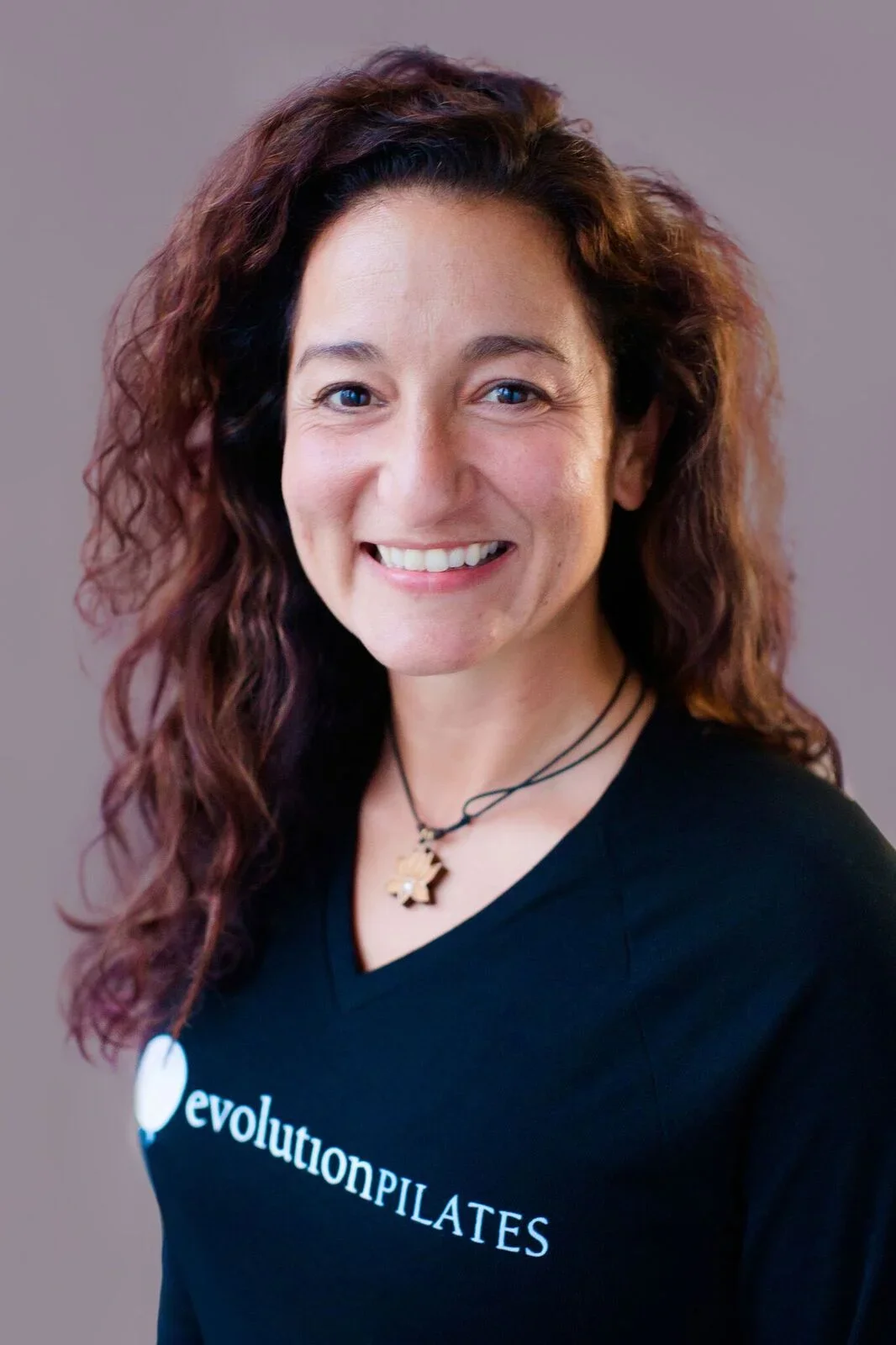 A woman with wavy, reddish-brown hair smiling at the camera, wearing a black t-shirt with white text that says 'evolutionPILATES', and a necklace with star-shaped pendants.