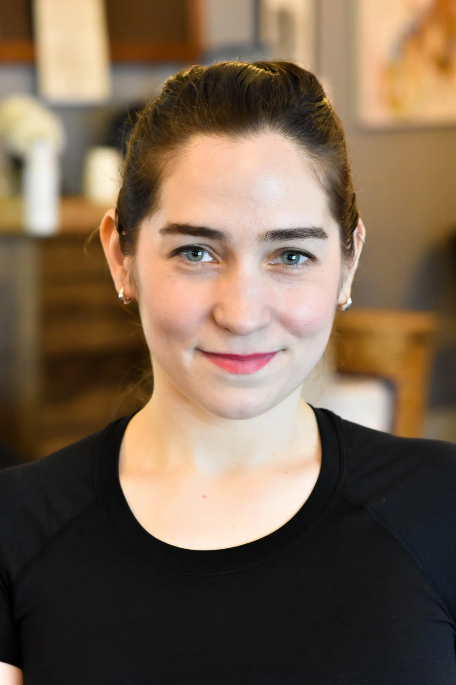 A young woman with brown hair pulled back, wearing a black top, smiling in an indoor setting with blurred background