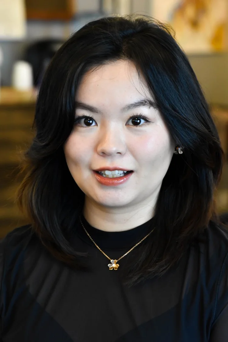 A woman with shoulder-length dark hair and light skin, smiling at the camera, wearing a black top, a gold necklace with a flower pendant, and earrings, with a blurred indoor background.