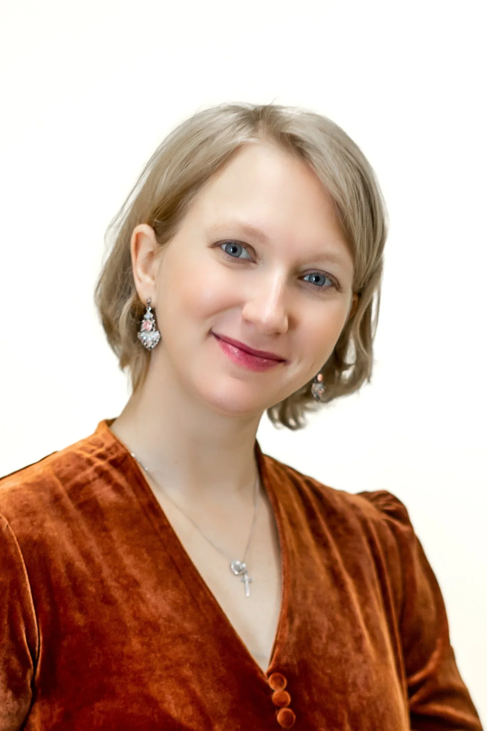 A woman with blonde hair and blue eyes wearing a rust-colored velvet top, earrings, and a necklace, smiling at the camera against a plain white background.