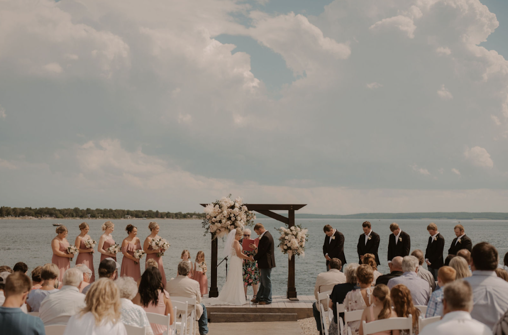 Outdoor wedding ceremony by the water with the bride, groom, officiant, bridesmaids, and groomsmen under a floral arch.