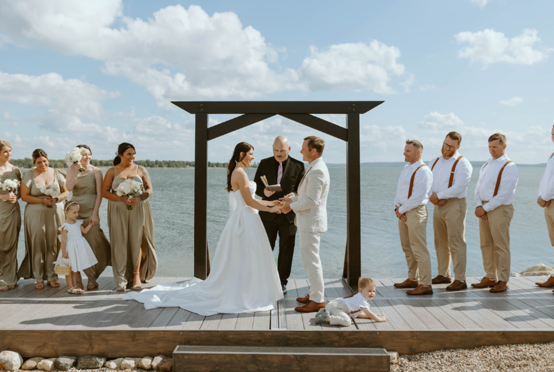 A wedding ceremony taking place outdoors on a dock over water, with a bride and groom exchanging vows under a black wooden arch, surrounded by bridesmaids, groomsmen, and a child, with cloudy sky above.