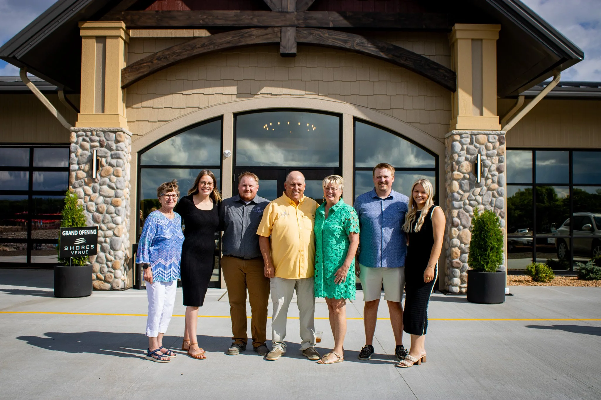 A group of seven people standing outside in front of a building with large glass windows and stone pillars, posing for a photo on a sunny day during a grand opening event.