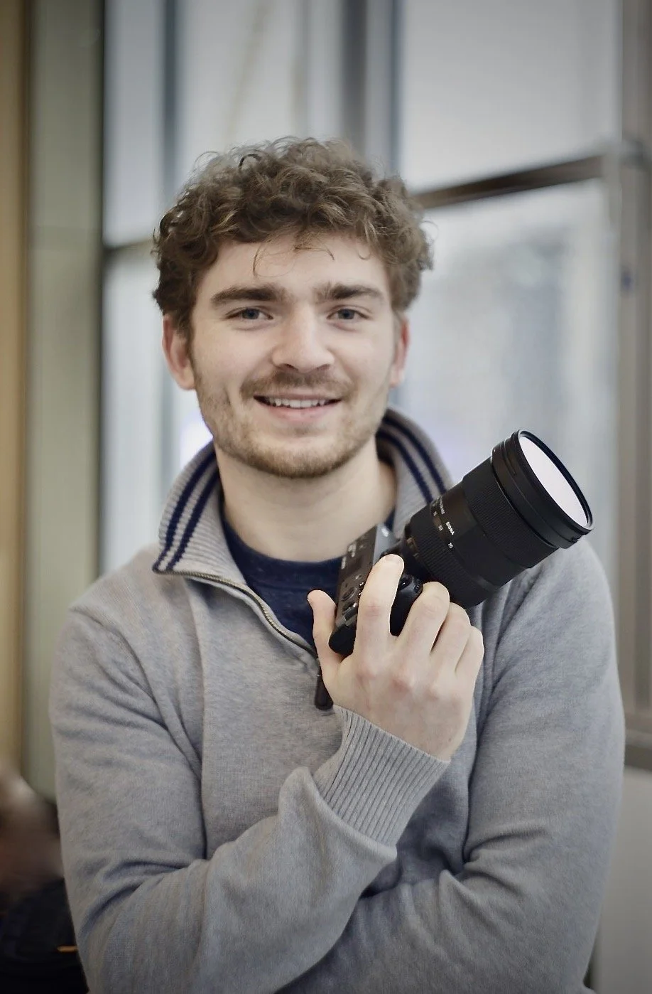 A young man with curly hair smiling and holding a professional camera with a large lens inside a room with windows.