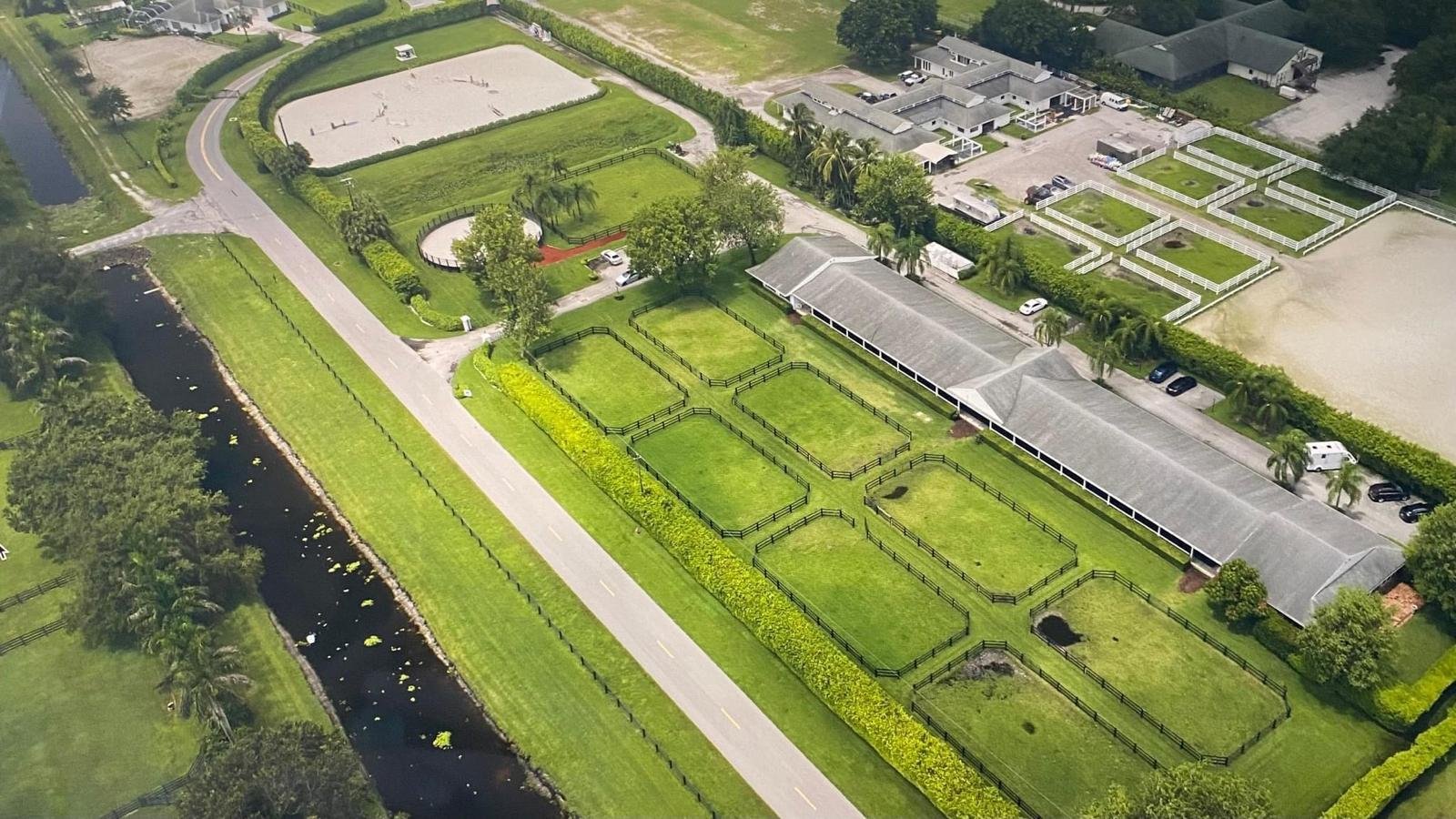 Aerial view of a horse farm with paddocks, training arenas, and buildings surrounded by greenery.
