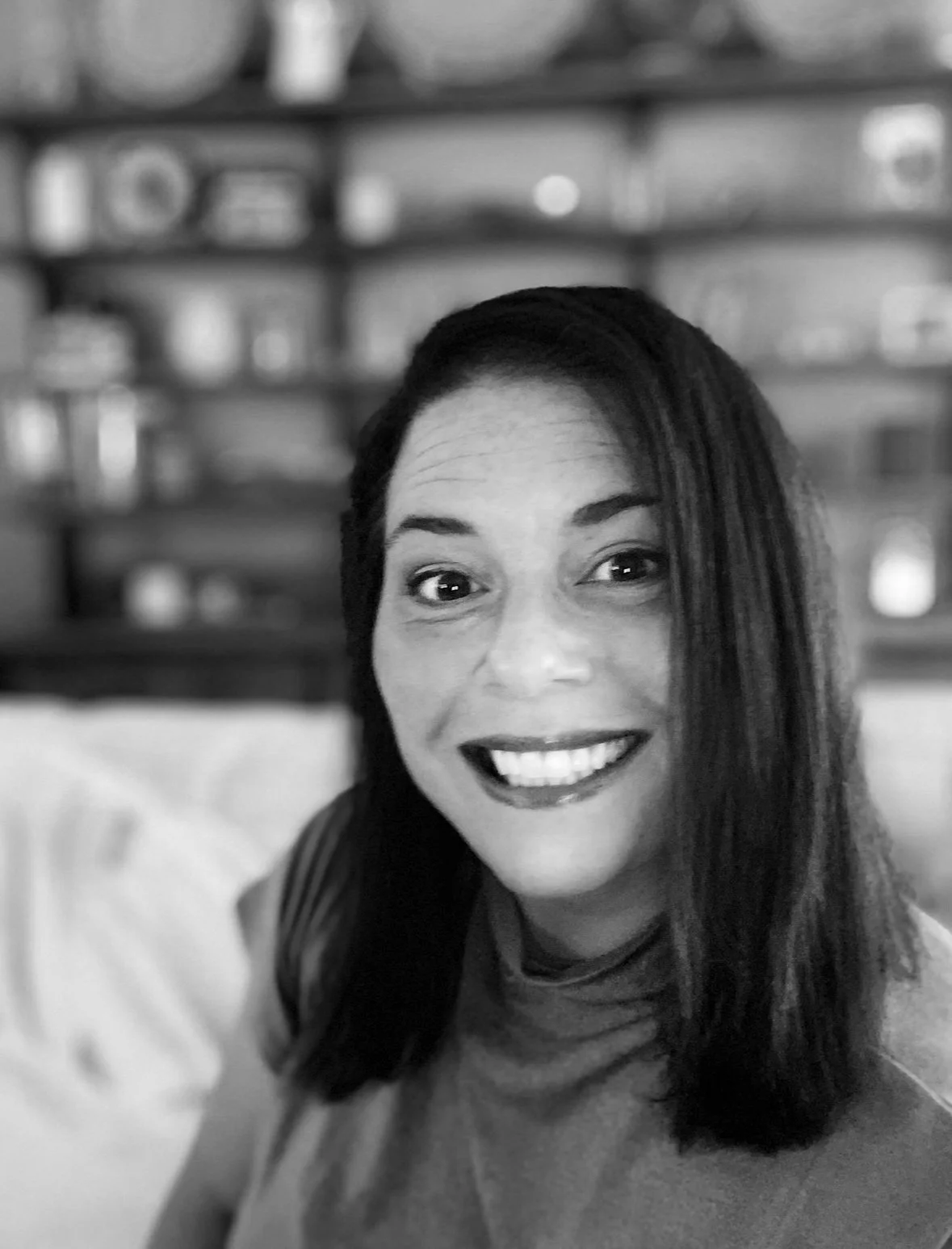 Black and white photo of a smiling woman with shoulder-length dark hair, in a room with shelves and various household items in the background.
