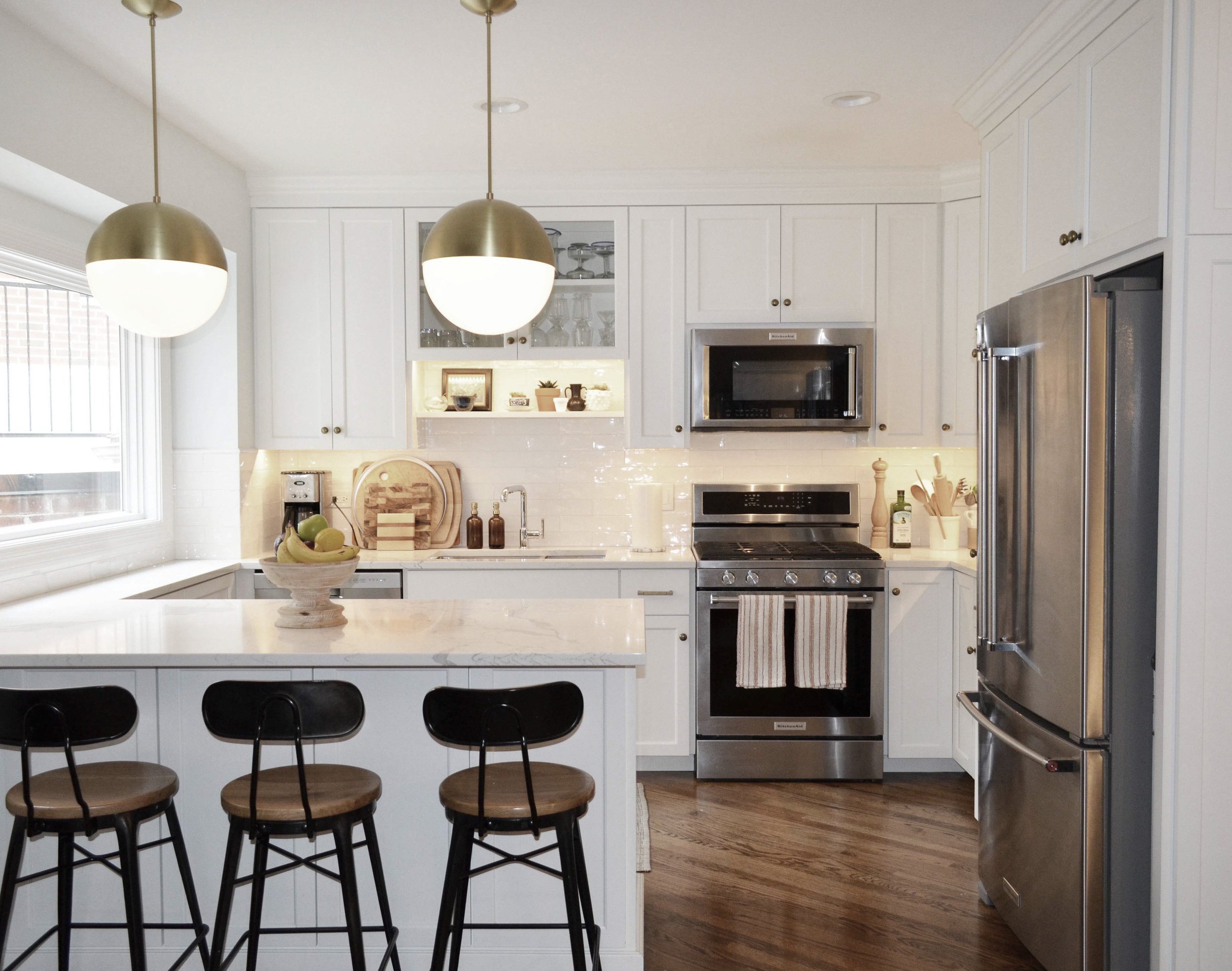 Modern kitchen with white cabinets, stainless steel appliances, a marble countertop island, three black and wood bar stools, and pendant lighting.
